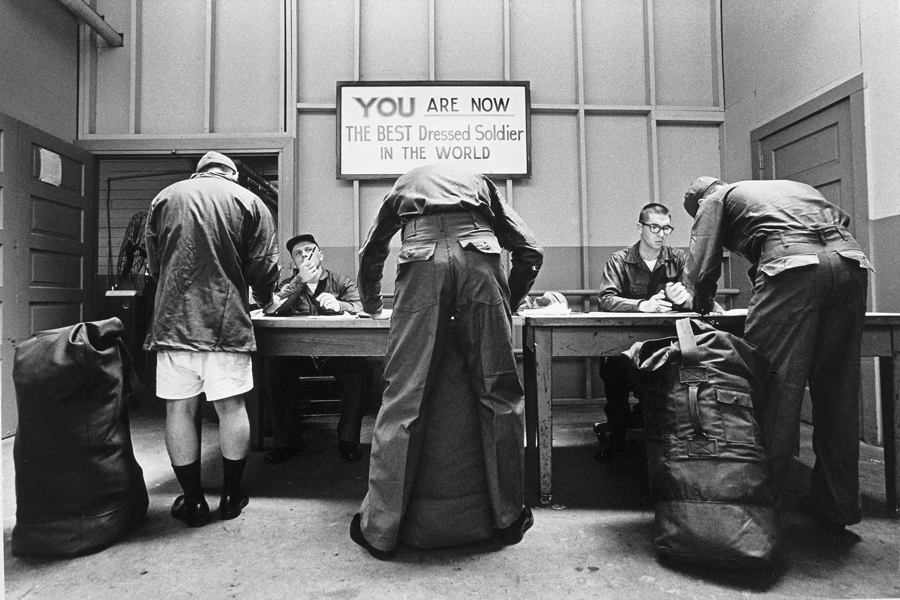 Three recently drafted soldiers sign papers under a sign that reads "You are now the best dressed soldier in the world."