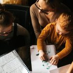 Little boy sits on his Mom's lap and draws while she helps older daughter with her schoolwork.