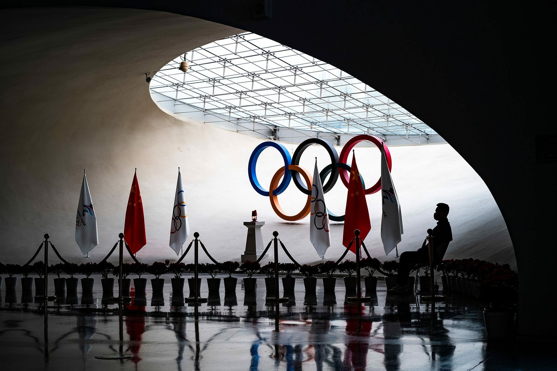 A man guards the olympic flame, which stands in front of Olympic rings.