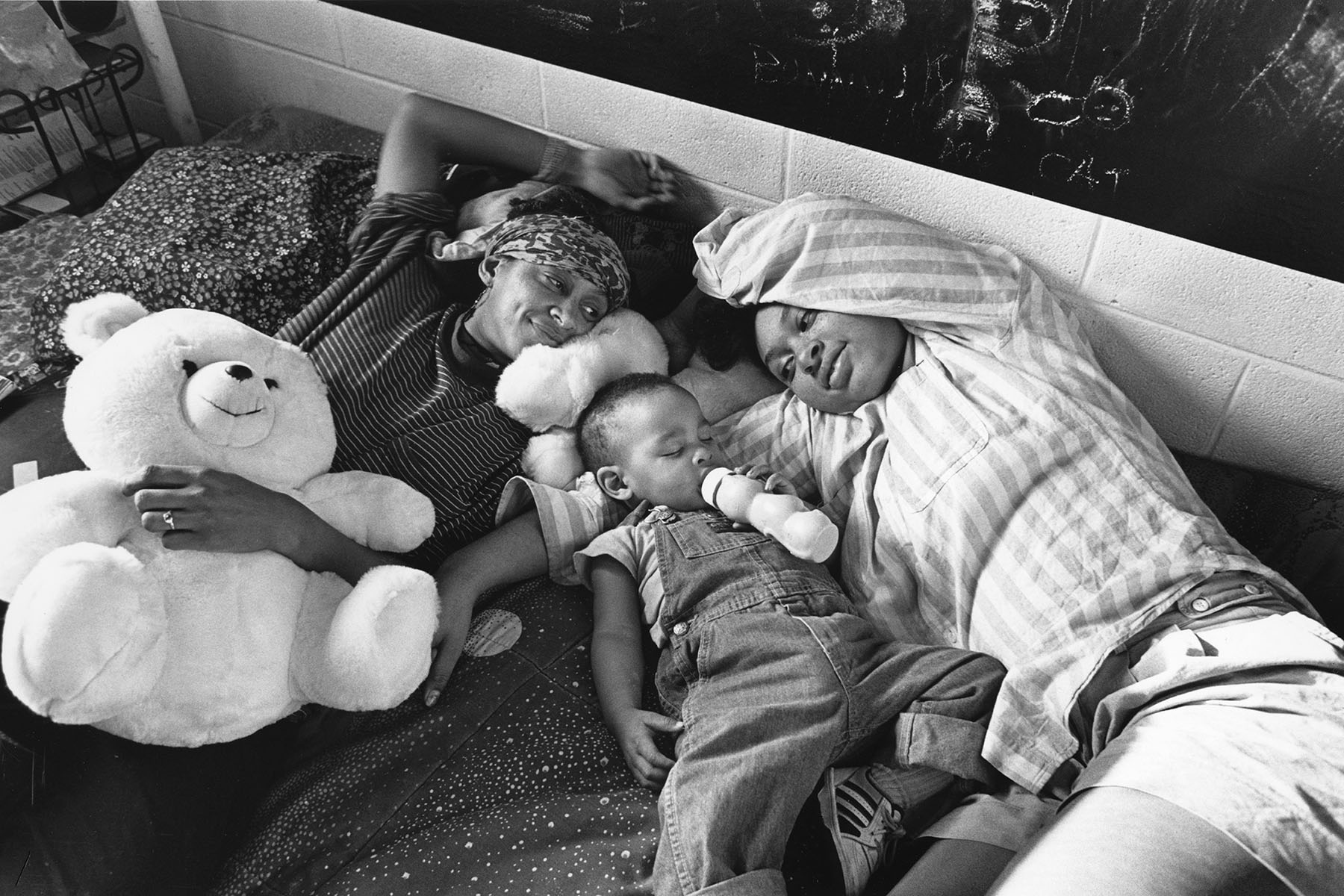 A grandmother, her daughter and her grandson lay on a bed together. The grandson is having his bottle and the grand mother is clutching a large teddy bear.
