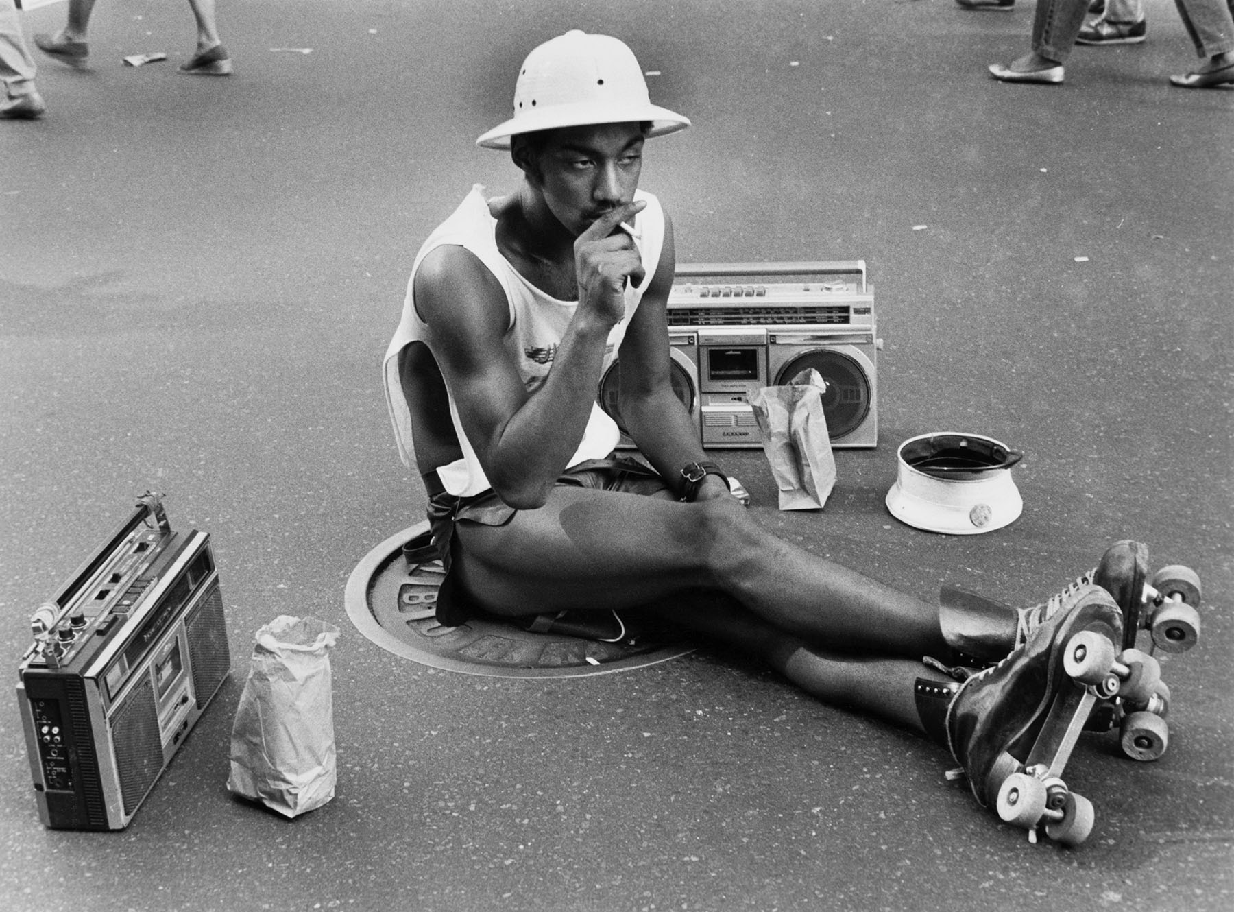 A young man wearing roller skates a pith helmet sits on the ground as he smokes a cigarette.