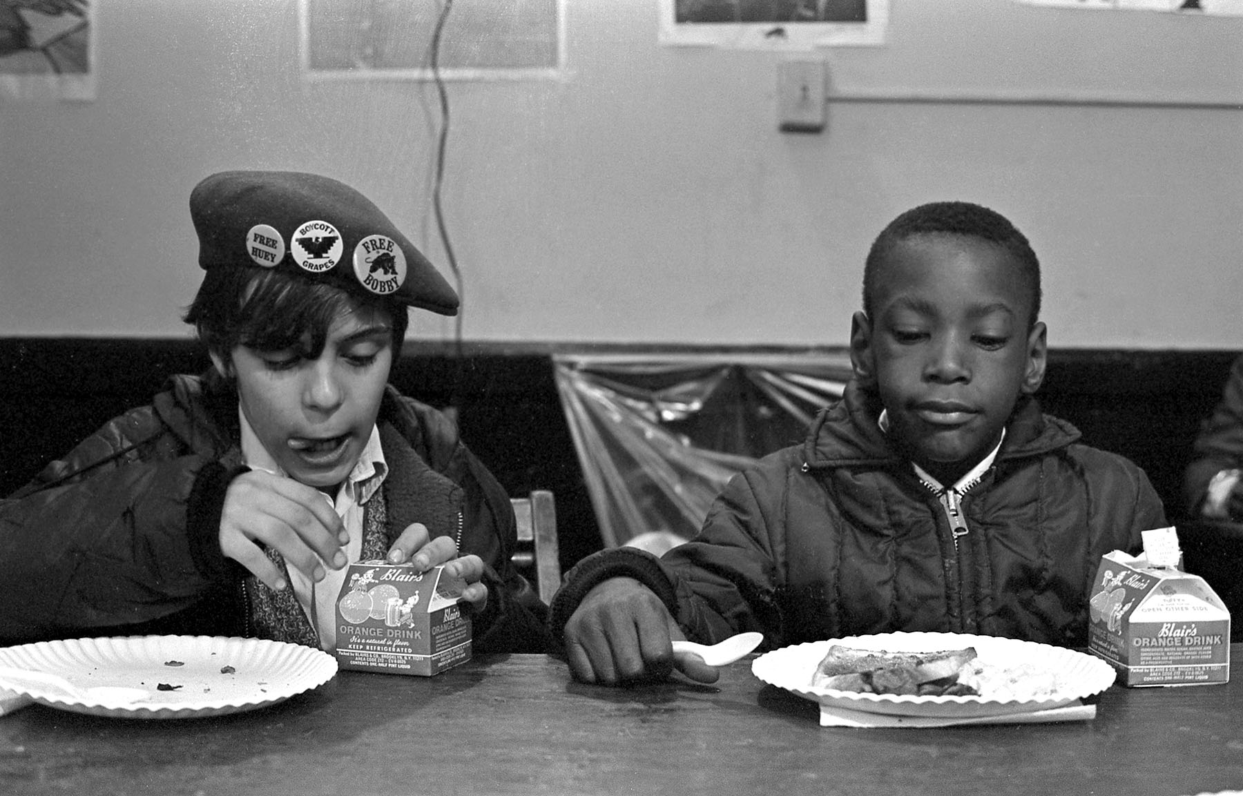 Two young boys seated at a table eat breakfast. The boy on the left wears a beret with buttons that read 