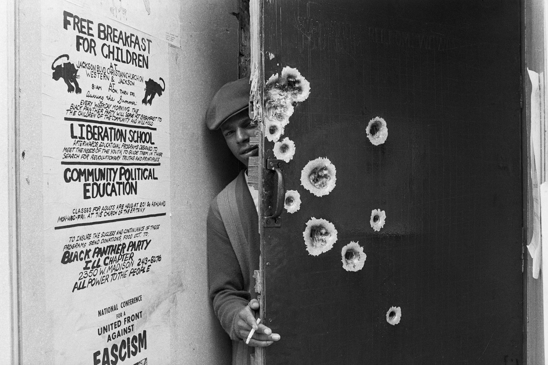A man peeks his head through a bullet pocked door. A poster near him advertises 
