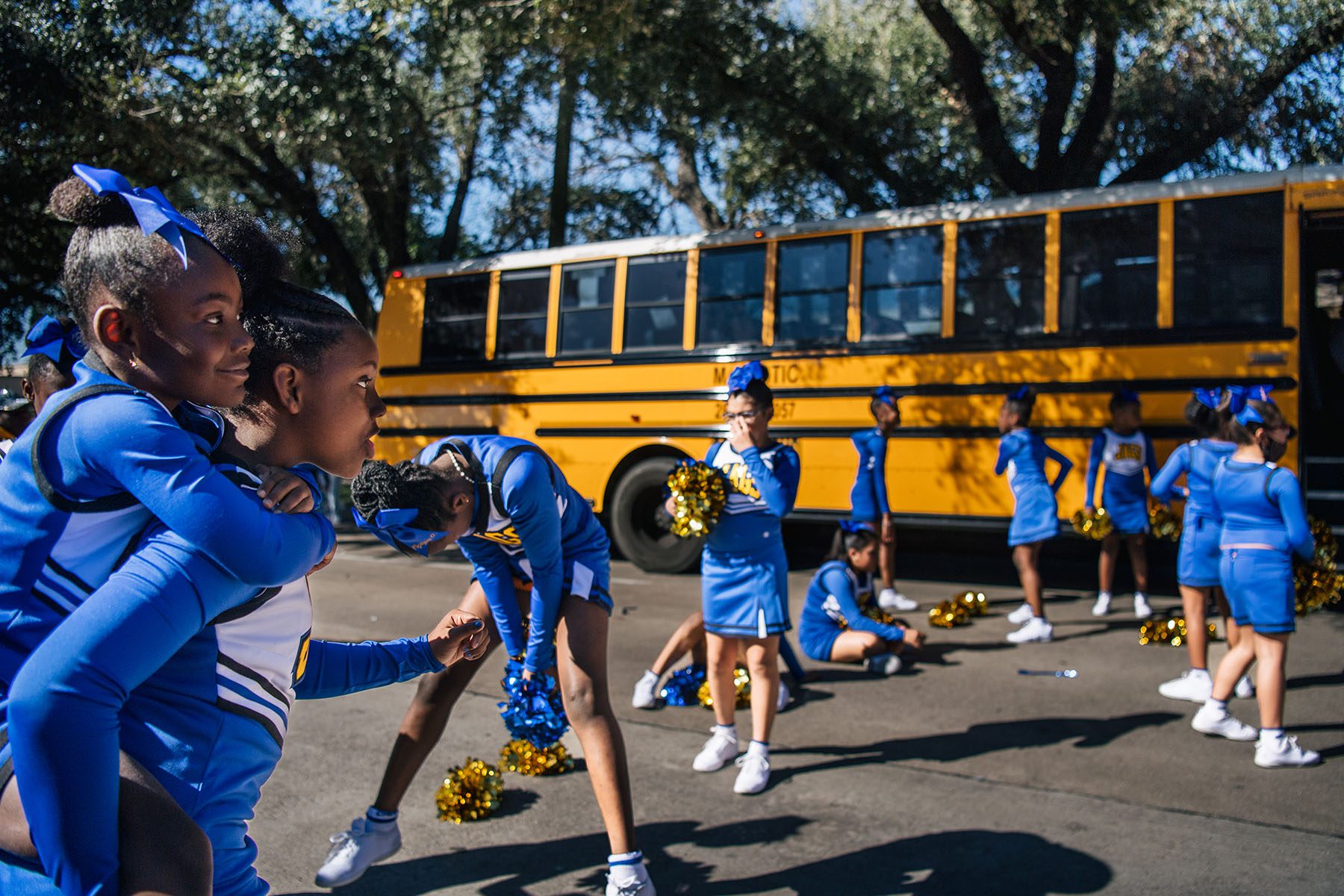 Young cheerleaders in blue uniforms, ribbons and golden pom-poms stretch and play before boarding a school bus.