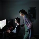 A mother stands over her son while he works on a computer.