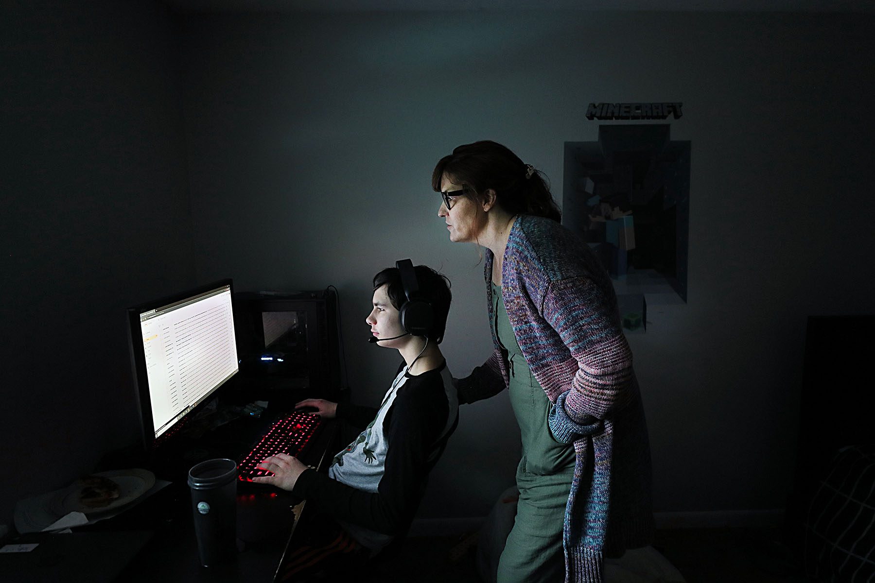 A mother stands over her son while he works on a computer.