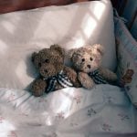 Two teddybears tucked in to a toddler's crib with a crib bumper.