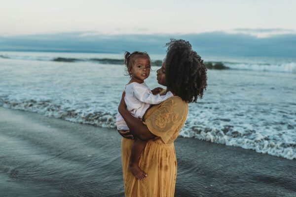 A pregnant mom and her daughter embrace near the ocean.