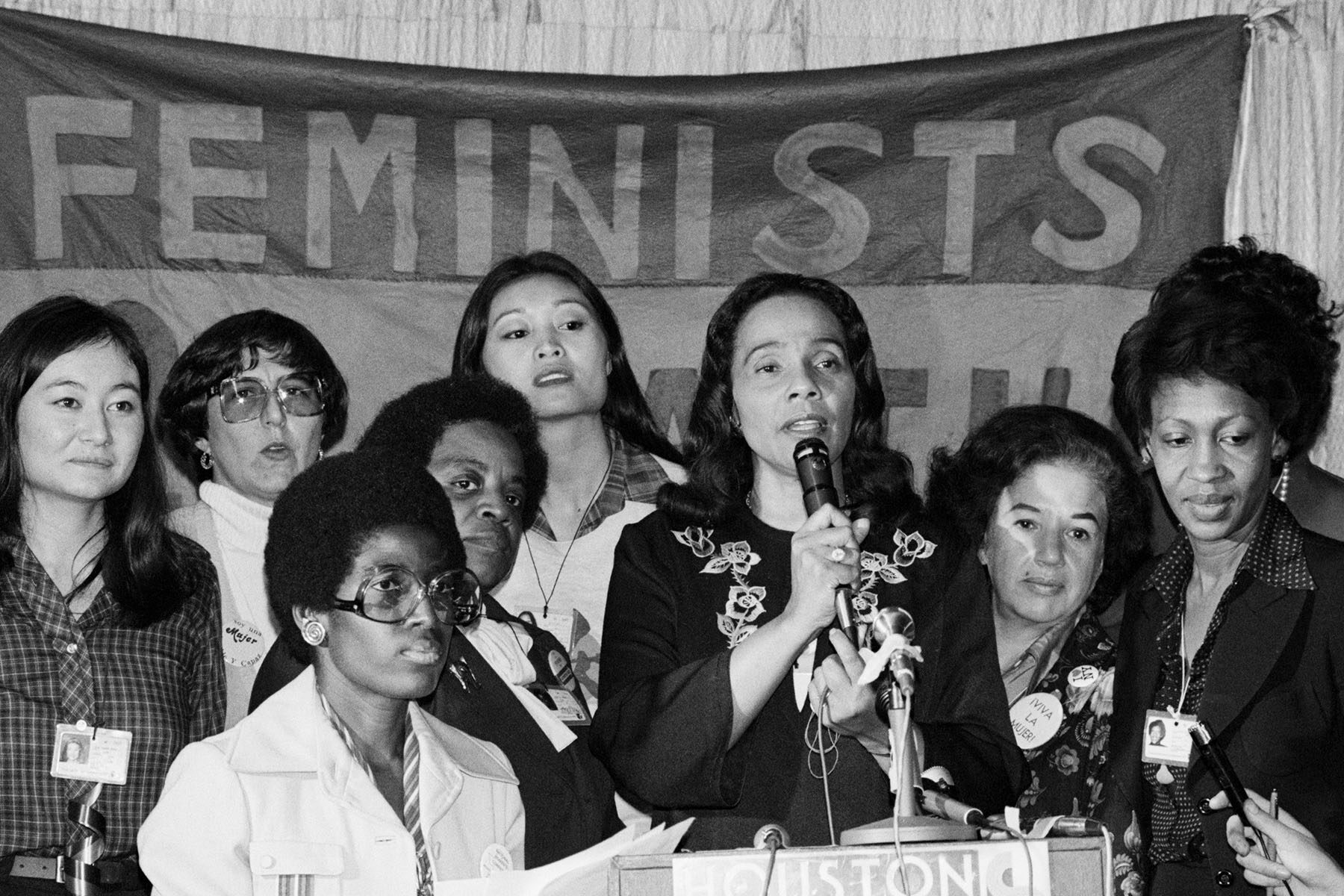Surrounded by women, Coretta Scott King speaks at a podium during the National Women's Conference