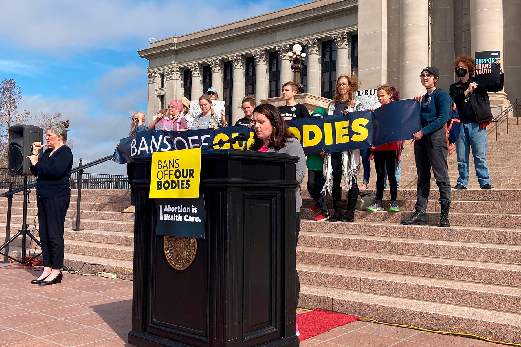 A woman speaks at a podium on which hangs a sign that reads "Bans off Our Bodies" and "Abortion is Health Care." People stand behind her holding a banner that reads "Bans off our bodies." A sign language interpreter signs next to the speaker.