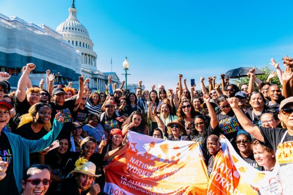 A large group of people hold their fists up in the air and smile in front of the Congress building. They are wearing t-shirts that read 