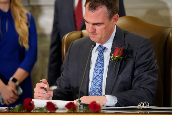 Kevin Stitt sits at a desk and signs into law a bill criminalizing abortions. He is wearing a red rose on his lapel. Several other red roses are seen on his desk.