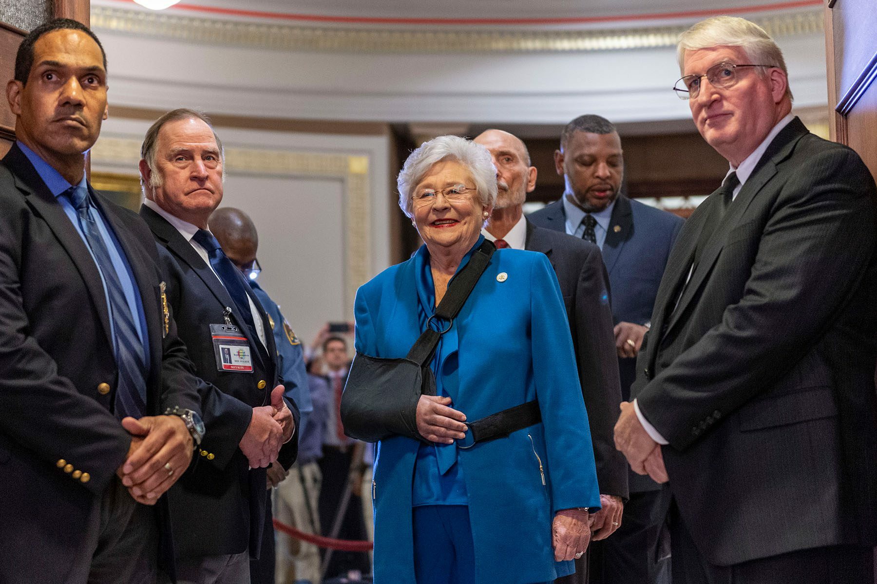 Gov. Kay Ivey wears a sling around her arm as she smiles before walking into the chamber. She is surrounded by several men.