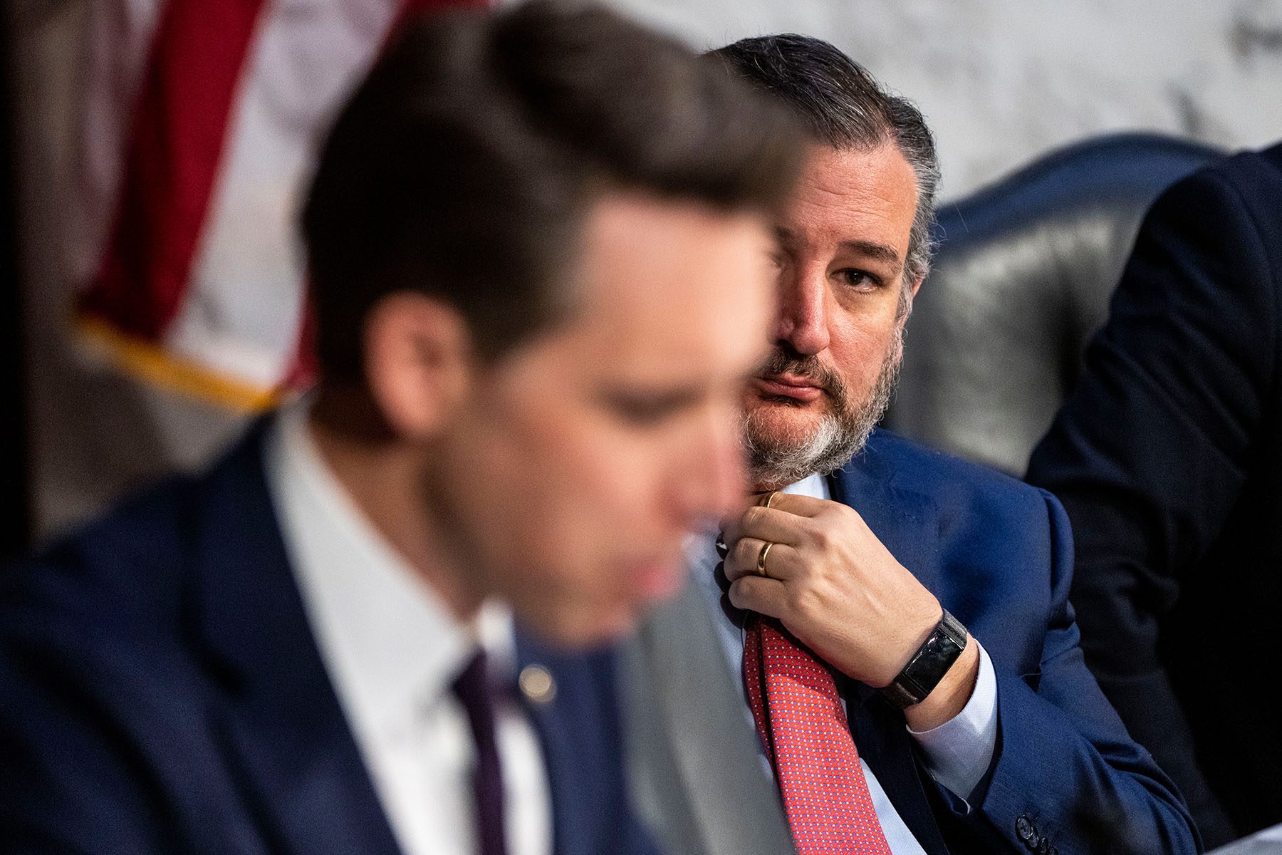 Sen. Ted Cruz adjusts his tie as Sen. Josh Hawley speaks during a meeting. Josh Hawley is blurred in the foreground, while Ted Cruz is in focus in the background.