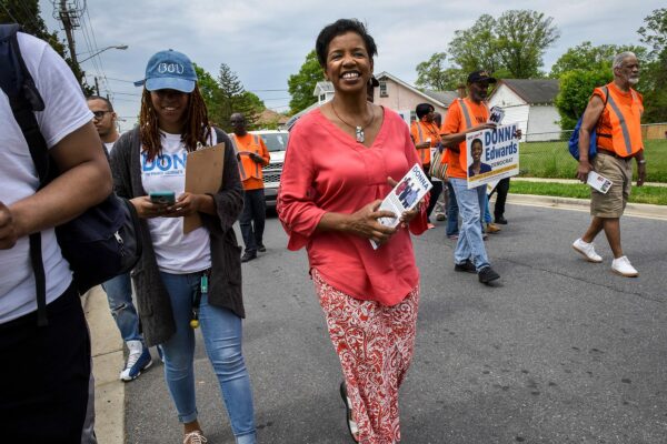 Donna Edwards walks down a street carrying a small pamphlet. Others around her carry signs that read 