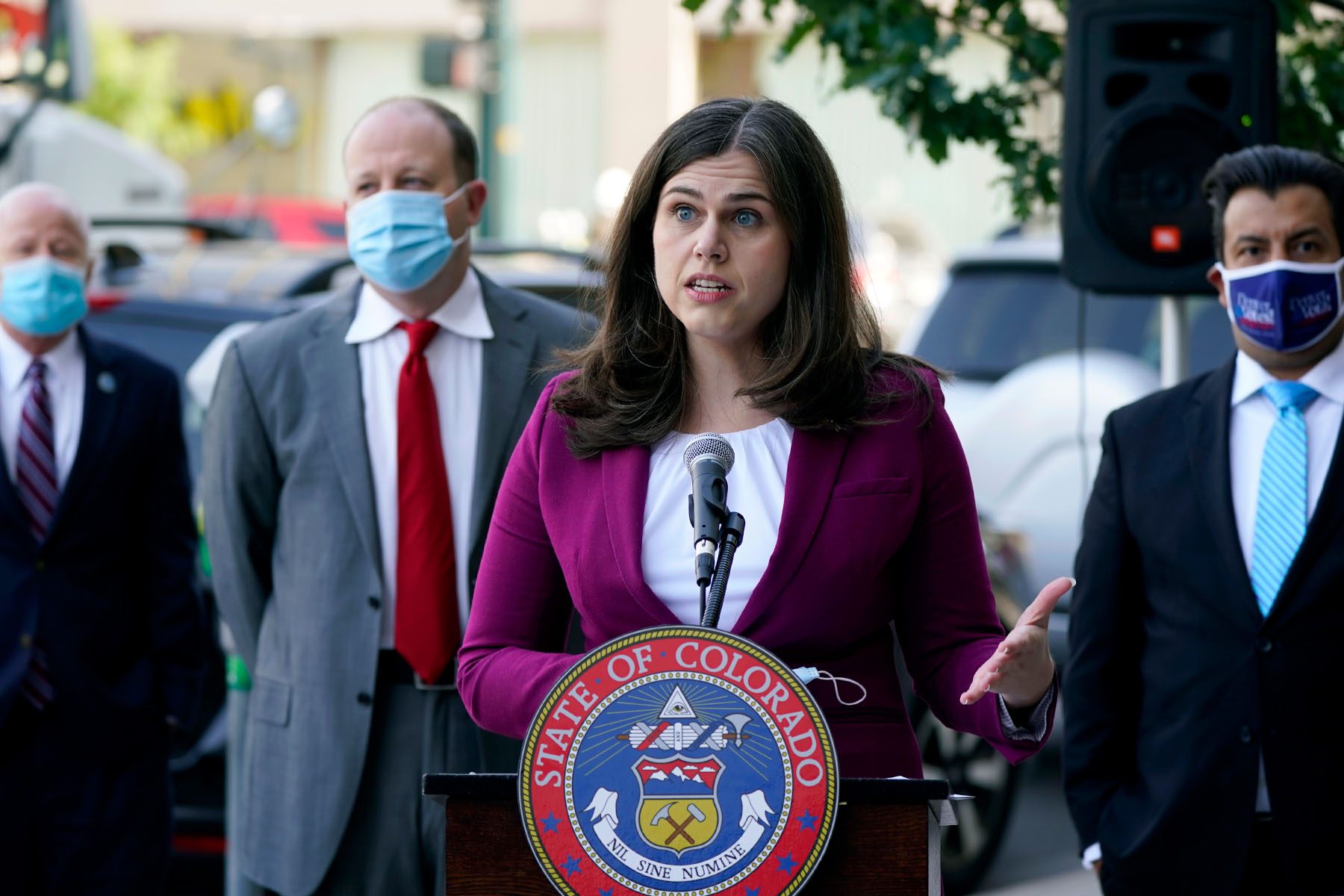 Jena Griswold speaks at a podium branded with the State of Colorado seal. Behind her are three state officials wearing face masks.