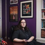 Julia Bascom poses in her DC office. There are pictures on the wall and knickknacks on the desk. Behind her two posters read 