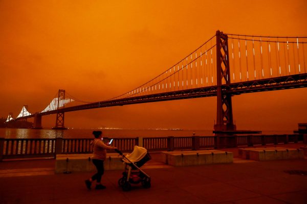 A woman pushing a stroller is seen near the Bay Bridge as a dark orange sky hands over downtown San Francisco.