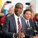 Dr. Joseph A. Ladapo is seen speaking at a podium. He is surrounded by people including Florida governor Ron DeSantis, who are blurred in the background.