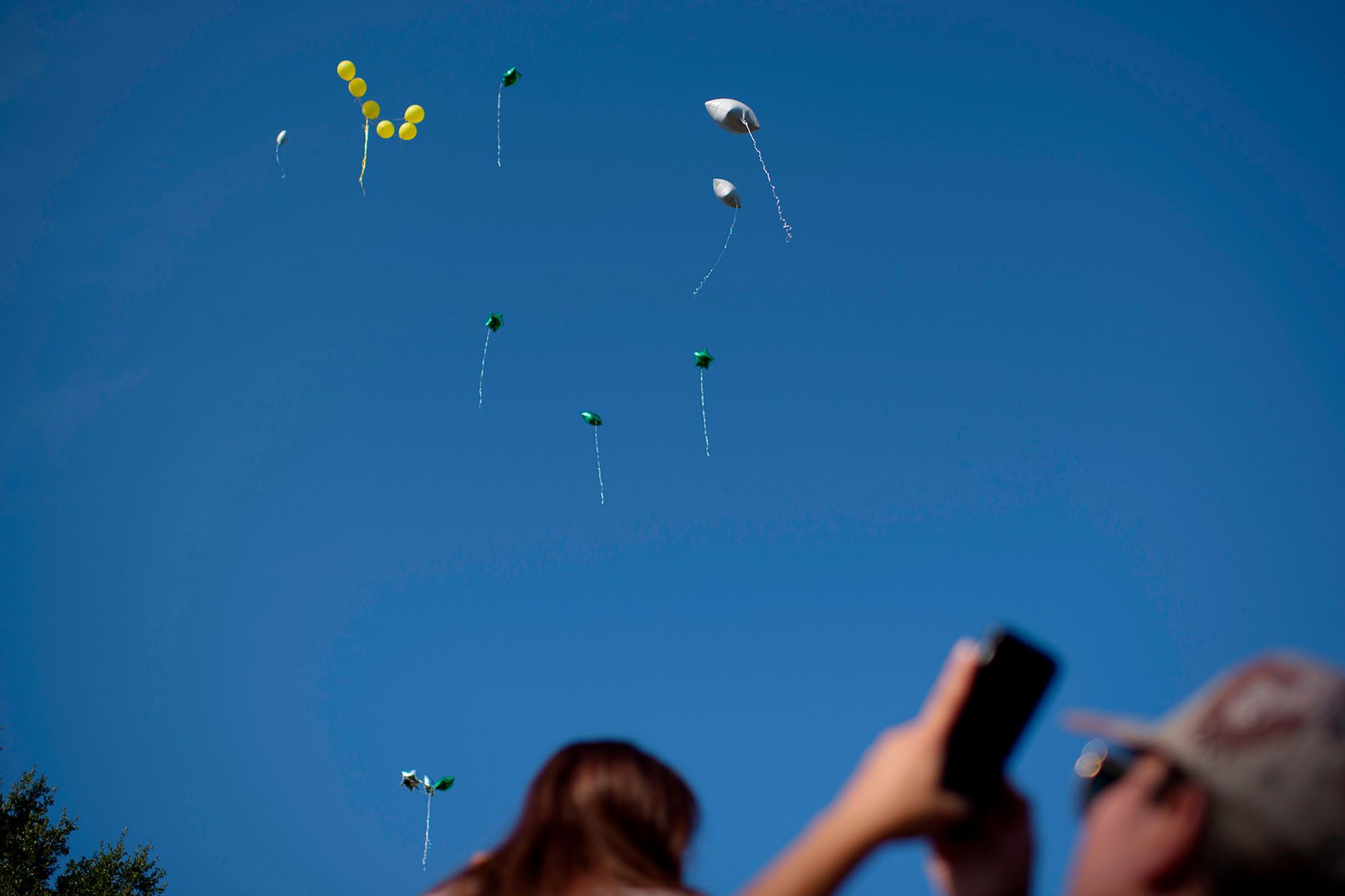 Star shaped balloons float away towards a blue sky as people look up and take photos on their cellphones.