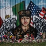 A mural depicts Vanessa Gullen in her military garb with angel wings. A Mexican flag and an American flag fly behind her. Candles, flowers are trinkets are scattered near the mural.