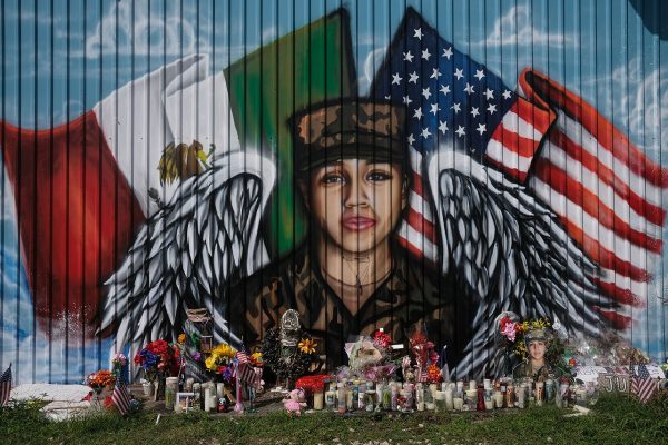 A mural depicts Vanessa Gullen in her military garb with angel wings. A Mexican flag and an American flag fly behind her. Candles, flowers are trinkets are scattered near the mural.