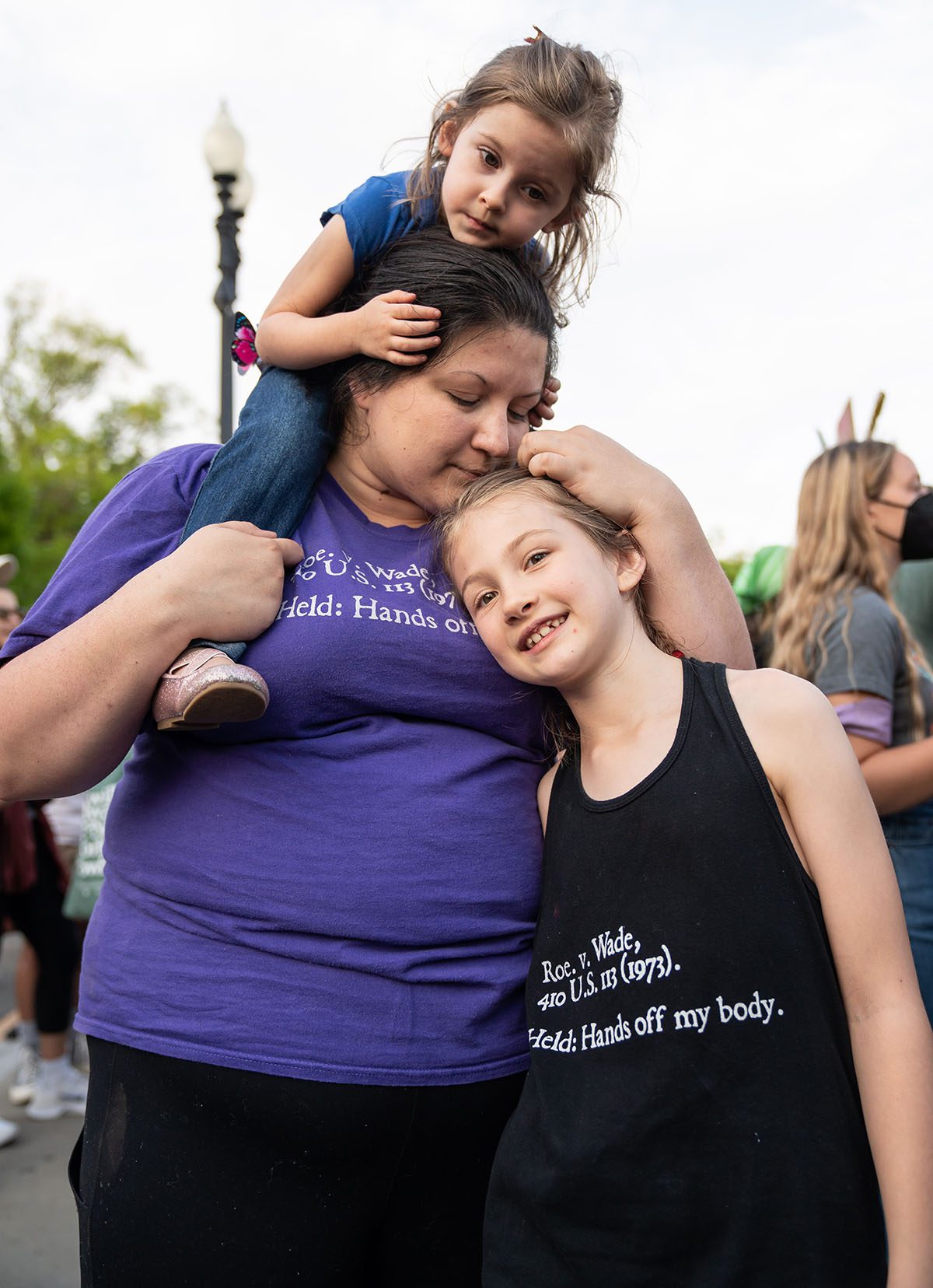 A mother hugs her daughter while holding the other on her shoulders. Mom and oldest daughter wear shirts that read "Roe v. Wade, 410 U.S. 113 (1973) Held: Hands off my body."