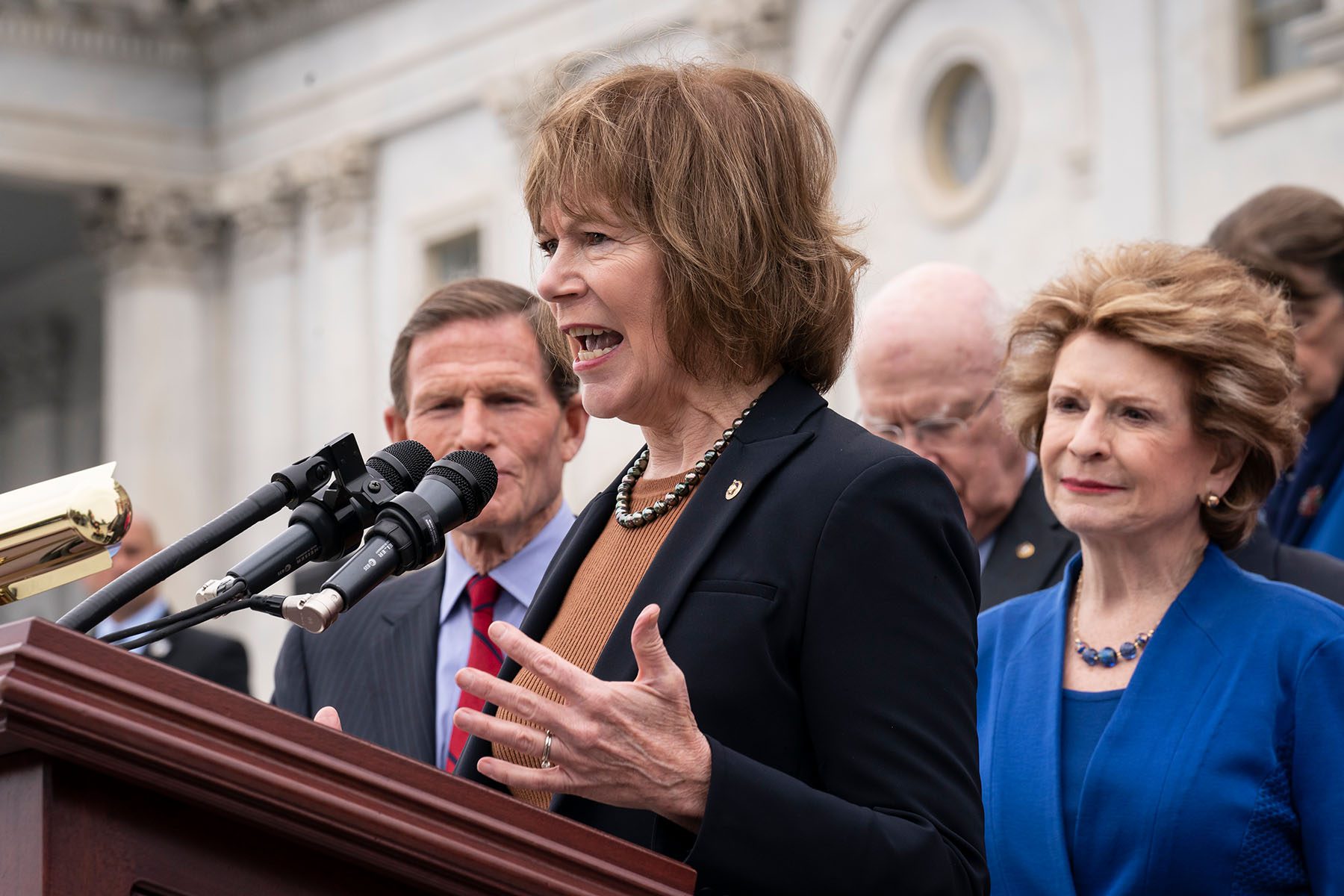 Tina Smith stands at a podium and speaks. She is surrounded by other members of congress.