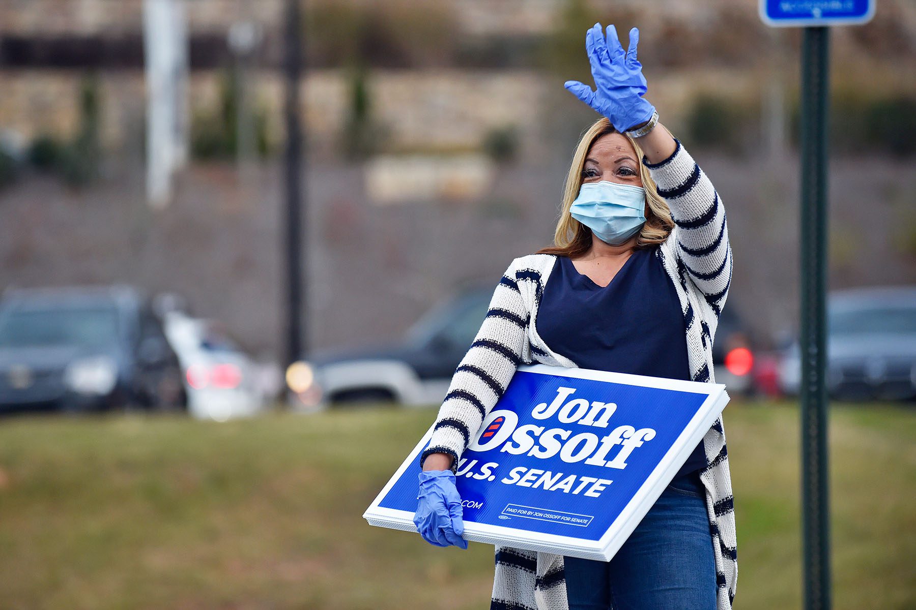 Lucy McBath waves at supporters of Jon Ossof. She is carrying yard signs that read "Jon Ossof U.S. Senate" and wearing a face mask and plastic gloves.