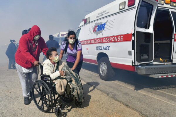 An elderly person in a wheelchair is transported to the back of an ambulance by two care workers as smoke is seen all around them.