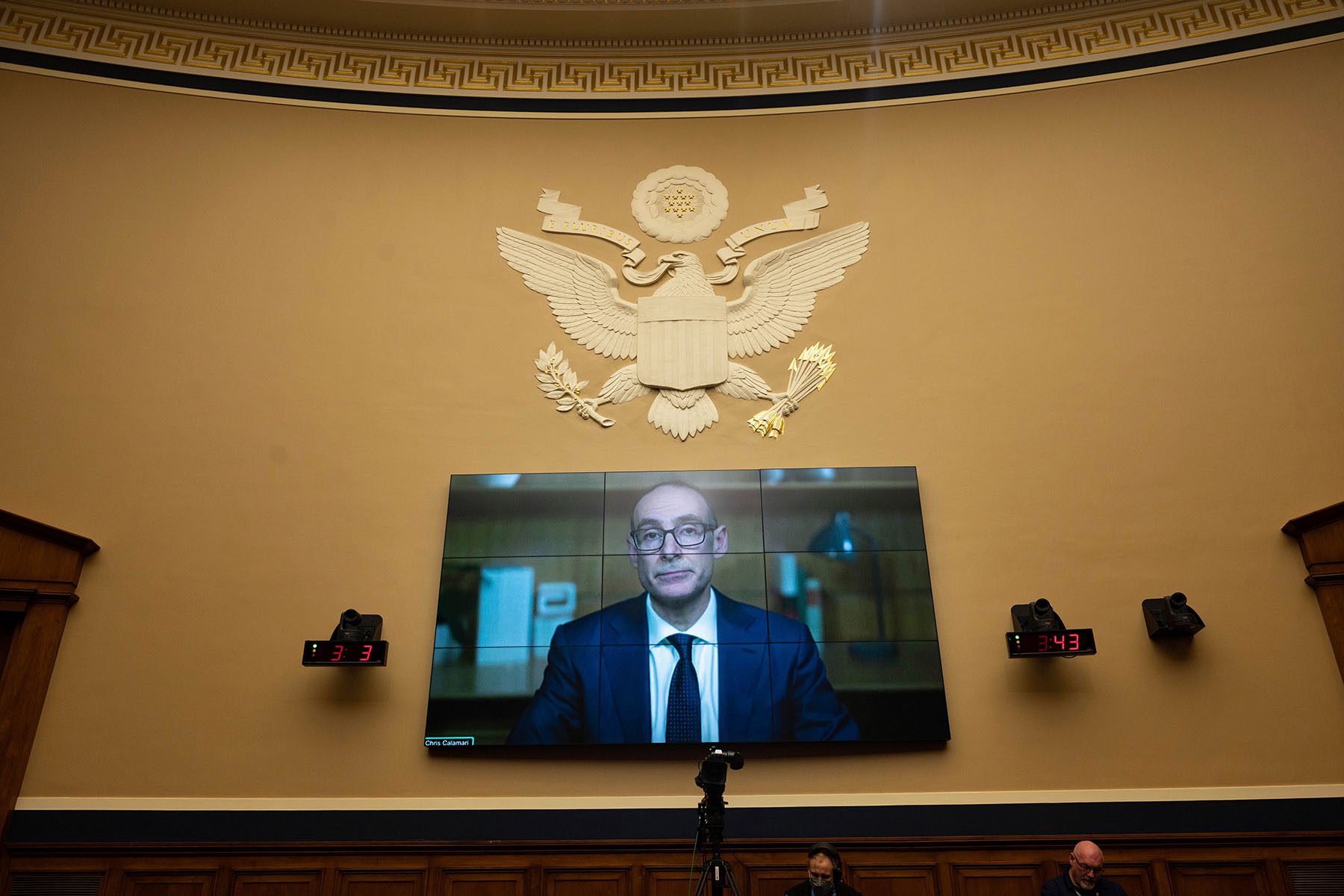 A television mounted on a wall in a Senate hearing room shows Abott Senior President Christopher Calamari as he testifies remotely.