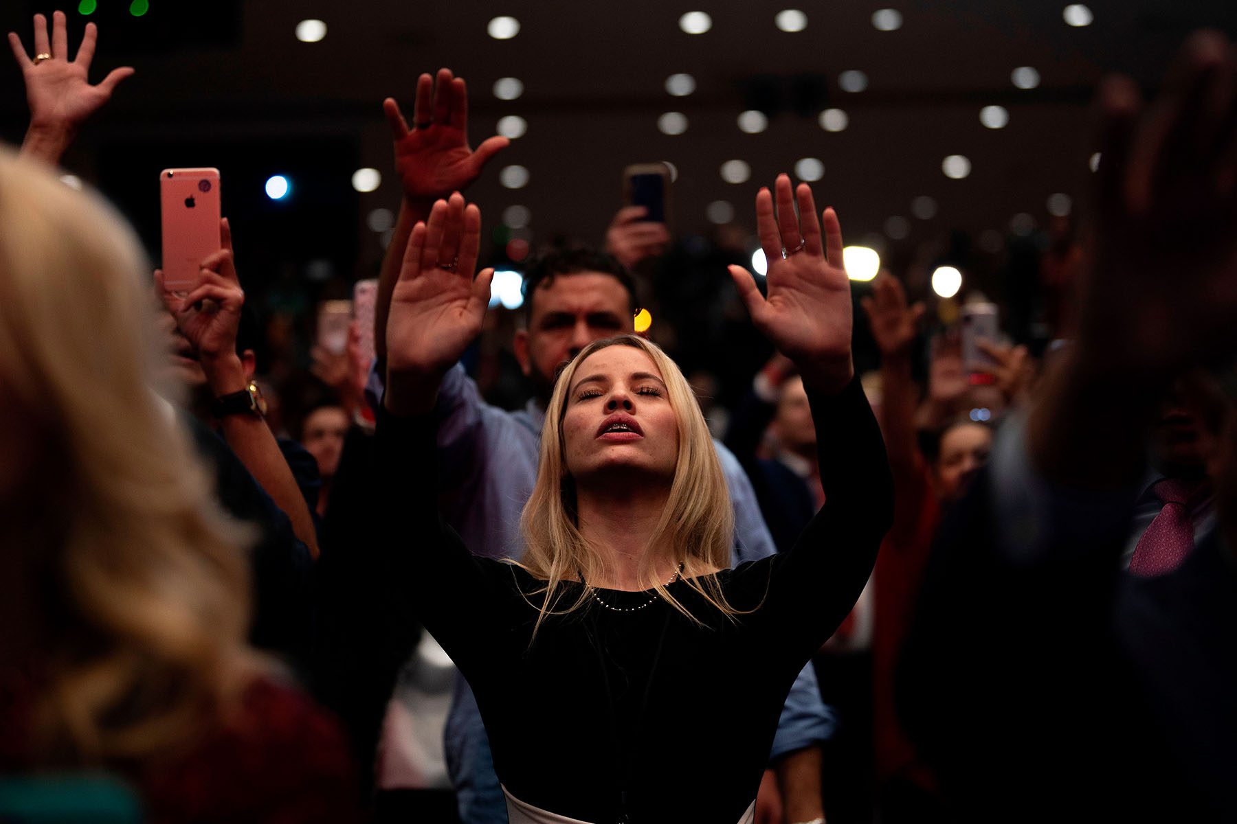 A woman raises both of her hands while she prays with her eyes shut. Around her, people raise their hands and take pictures on their cell phones.