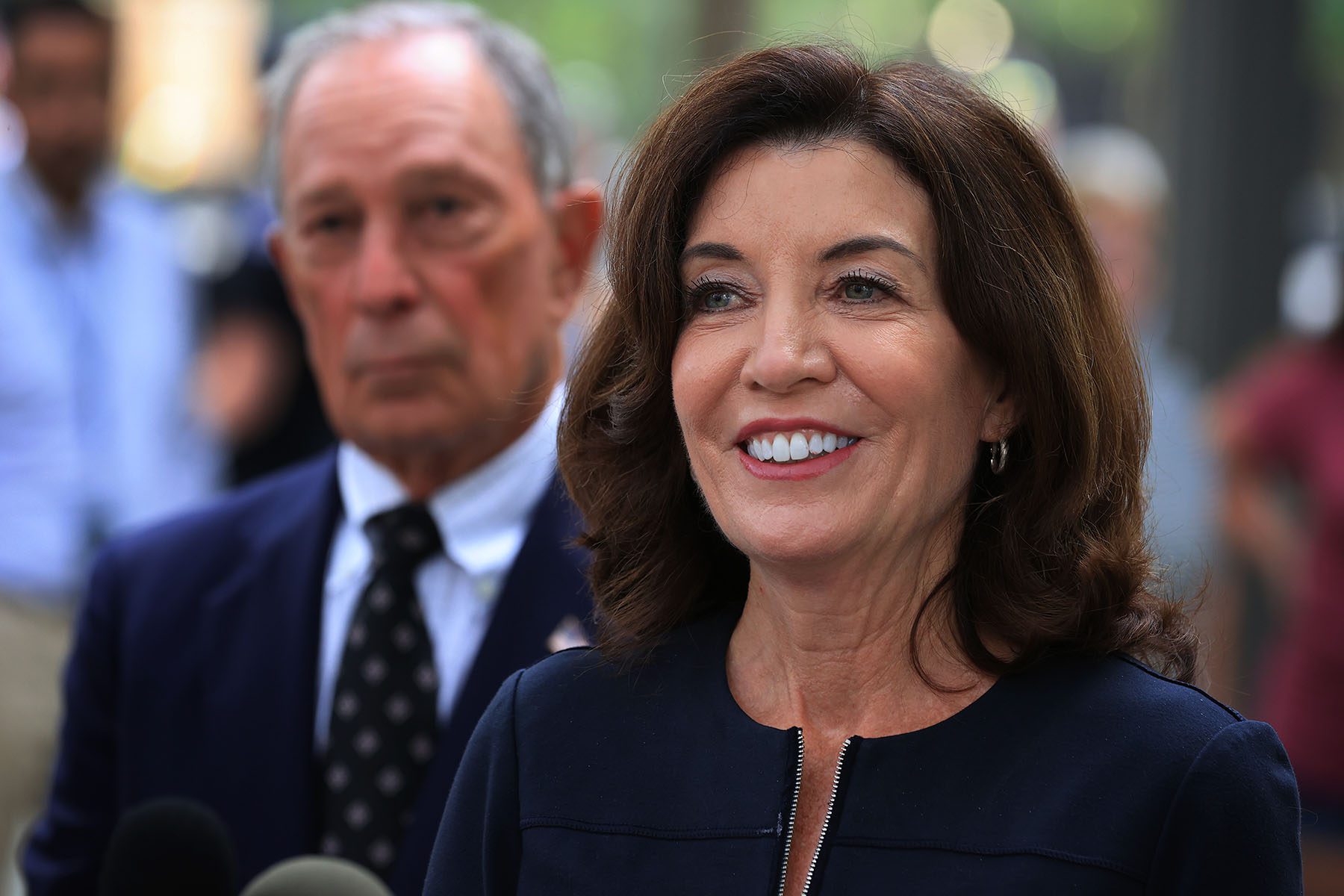 Kathy Hochul talks with reporters. Behind her is former New York City Mayor Mike Bloomberg.