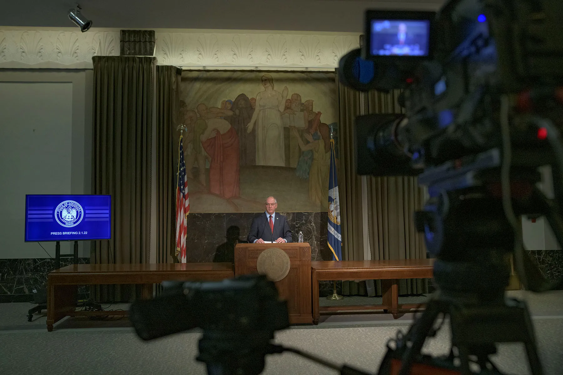 Louisiana Governor John Bel Edwards speaks during a press briefing. He is standing at a podium in the background of the image. In the foreground, a camera is seen recording the briefing.