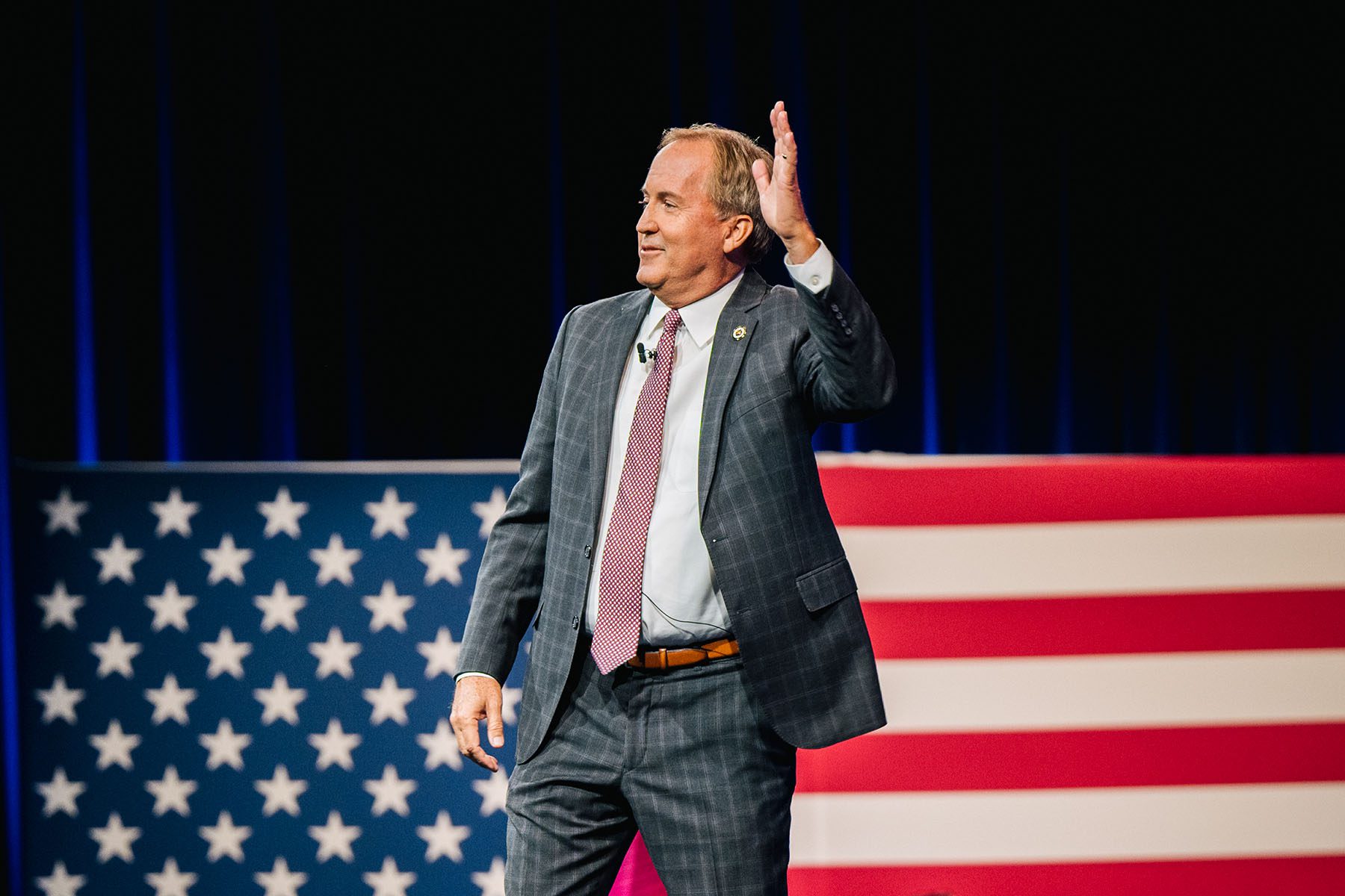 Ken Paxton waves after giving a speech. Behind him is a large American flag.