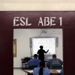 Prisoners are seen studying inside a class room. The teacher is resolving a math problem on the whiteboard. The door of the classroom reads 