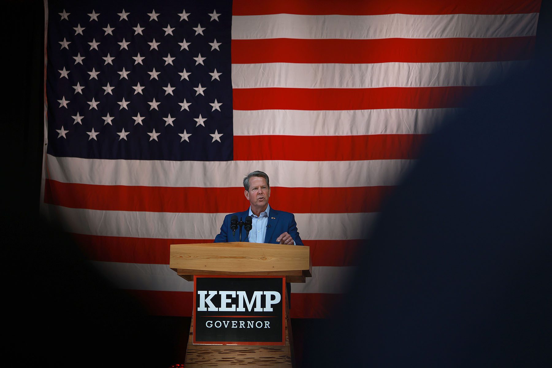 Brian Kemp speaks from a podium at a campaign event. The stage is adorned with a very large American Flag.