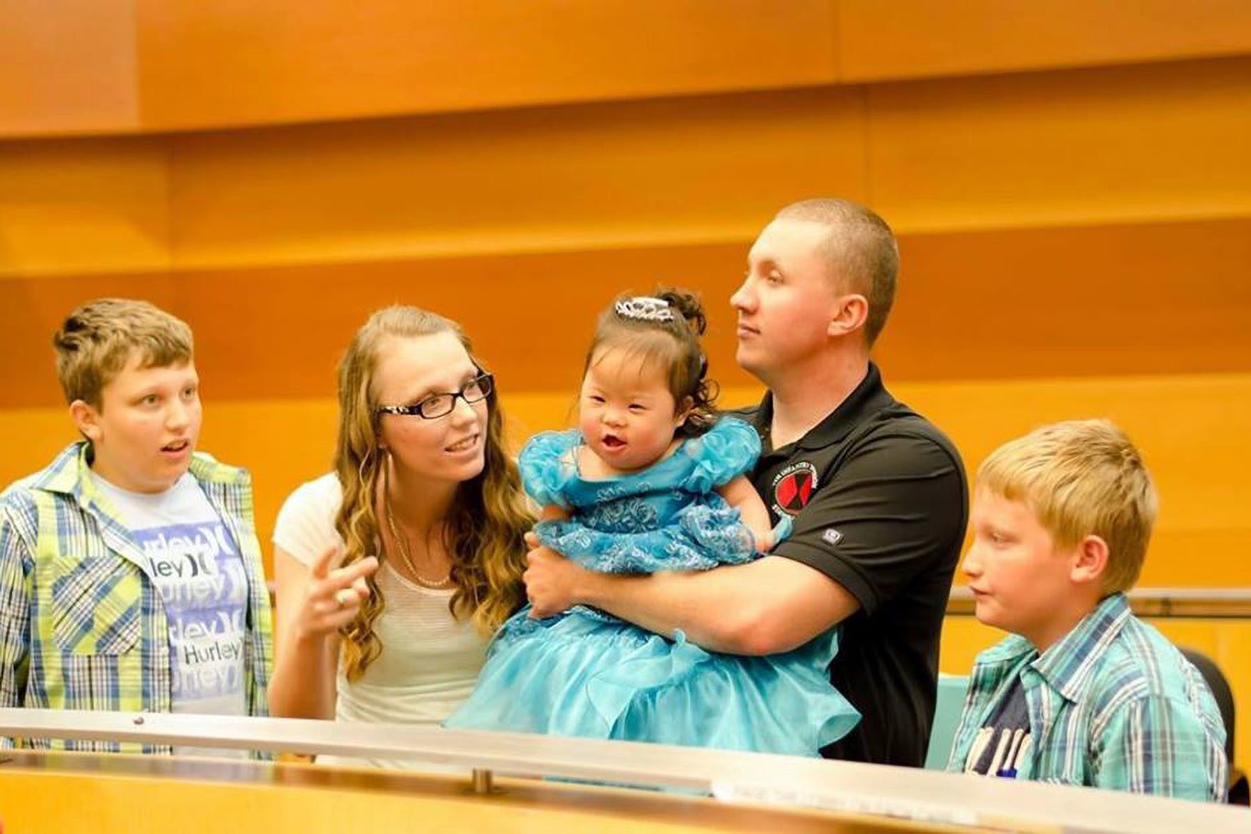 Austin Carrigg and her family are seen sitting down at adoption court. Carrigg's husband holds their young adopted daughter. The adopted daughter wears a princess dress and a small tiara and is smiling.