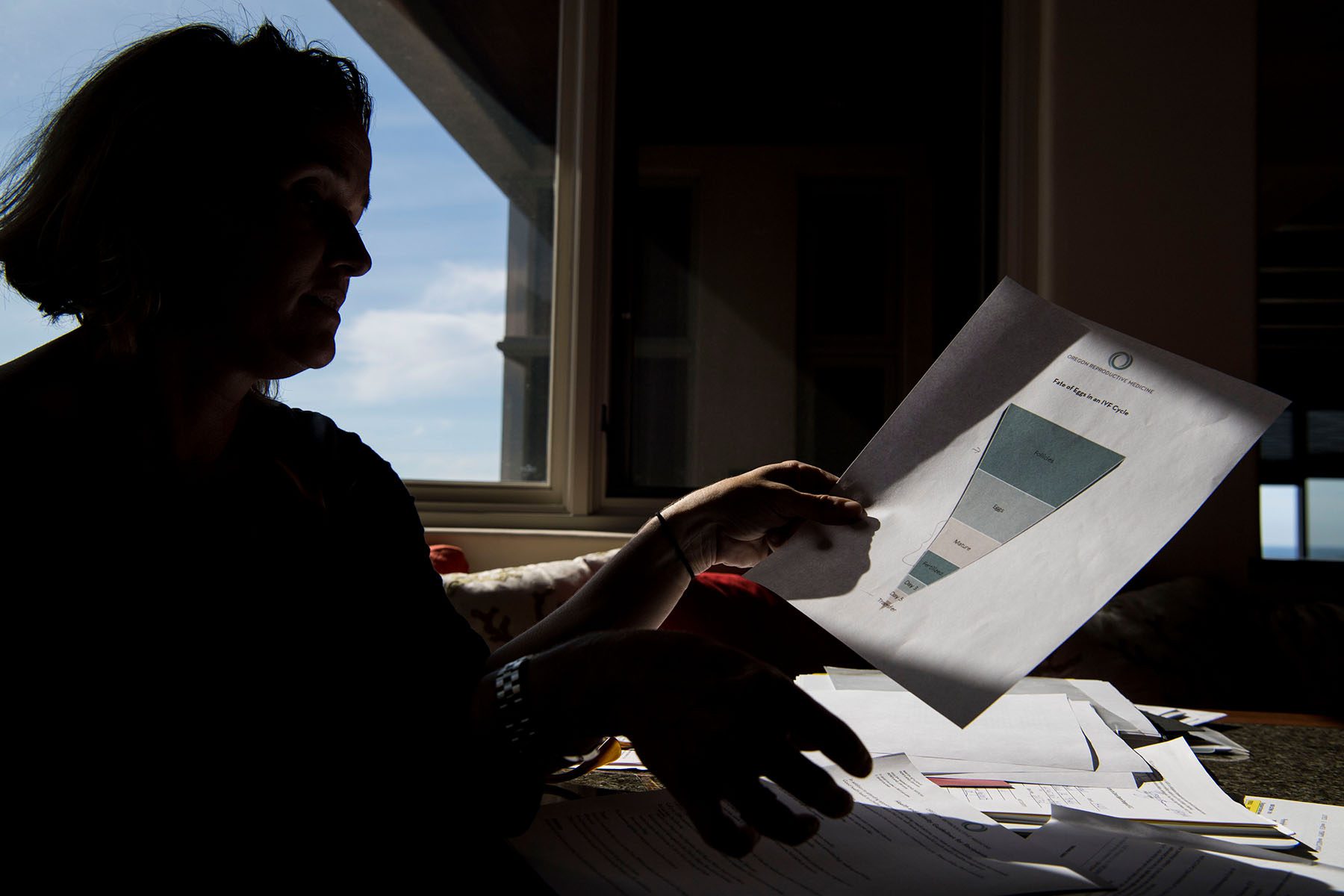 A woman goes through the paperwork for her egg freezing, IVF, and other fertility procedures and treatments. She is near a window and only her silhouette is visible while the sun shines on the paperwork she's reading.