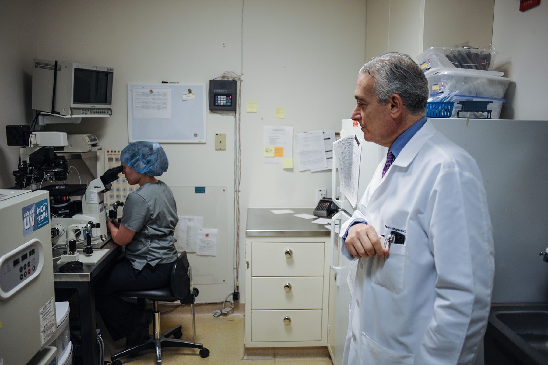 The Medical Director of Virginia Center for Reproductive Medicine watches an embryologist at work. She is looking through a microscope. He is standing a few feet back.