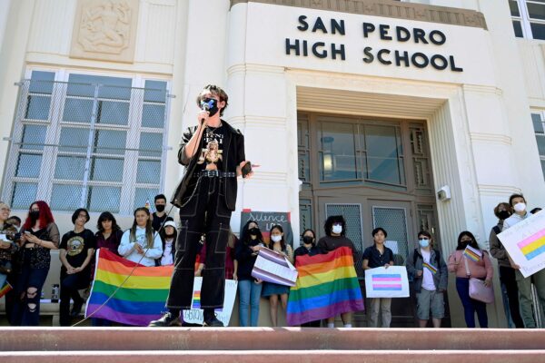 Dylan Keener holds a microphone while speaking in front of San Pedro High School. Behind him, students hold pride flag.