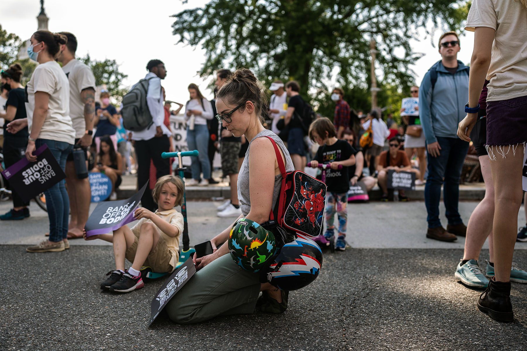 Emily Blake closes her eyes as she bows her head down while her two young sons play around her. She is carrying helmets and a spiderman lunchbox. One of her sons holds a signs that reads "Bans Off Our Bodies."