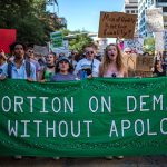 Protesters march while holding signs during an abortion-rights rally. A large banner reads 
