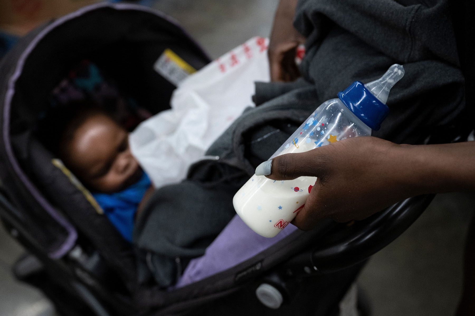 A half-full baby bottle is seen in a mother's hand as her baby sleeps in a stroller.