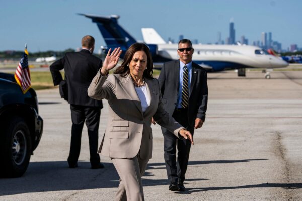Vice President Kamala Harris waves to the press before boarding Air Force Two. Behind her are two secret service agent and a plane.