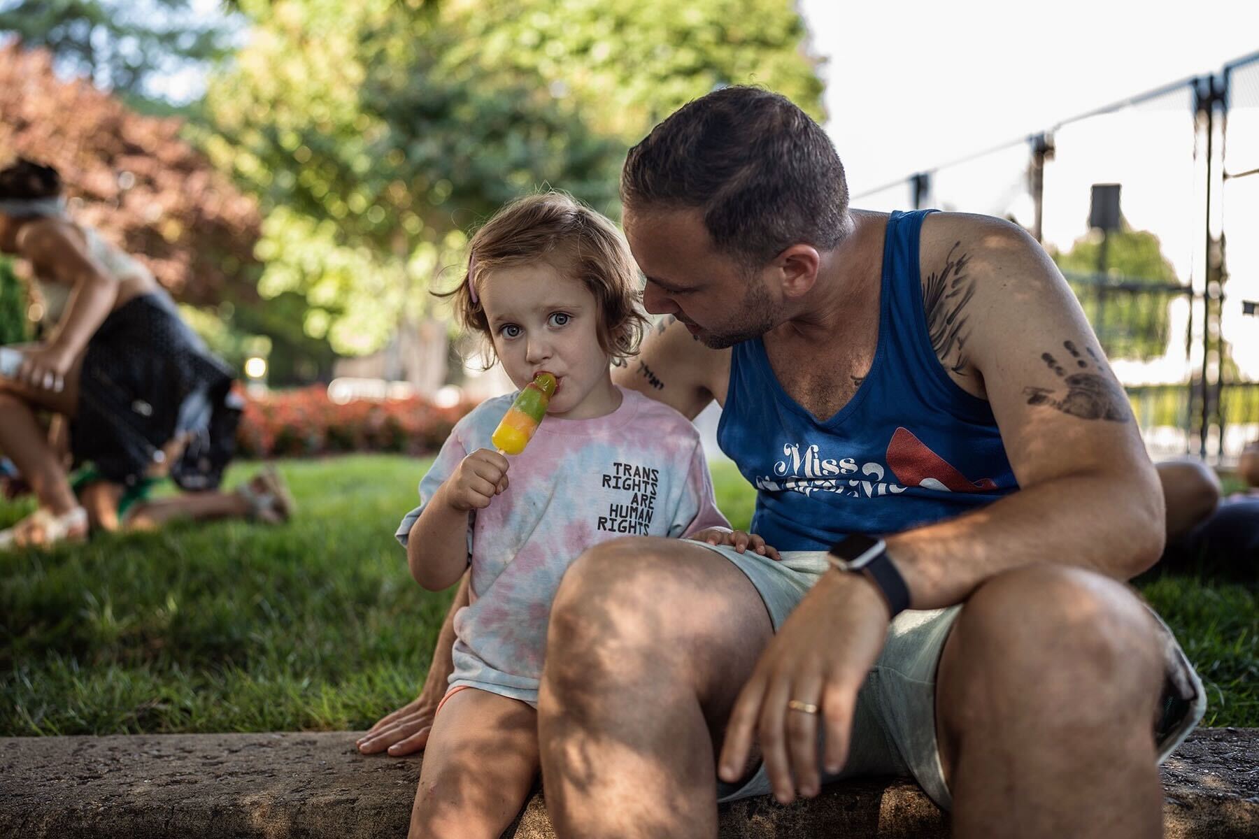 Eli Szenes-Strauss speaks to his young daughter as she eats a popsicle in the midst of a rally. Daughter Etta wears a shirt that reads "trans rights are human rights."