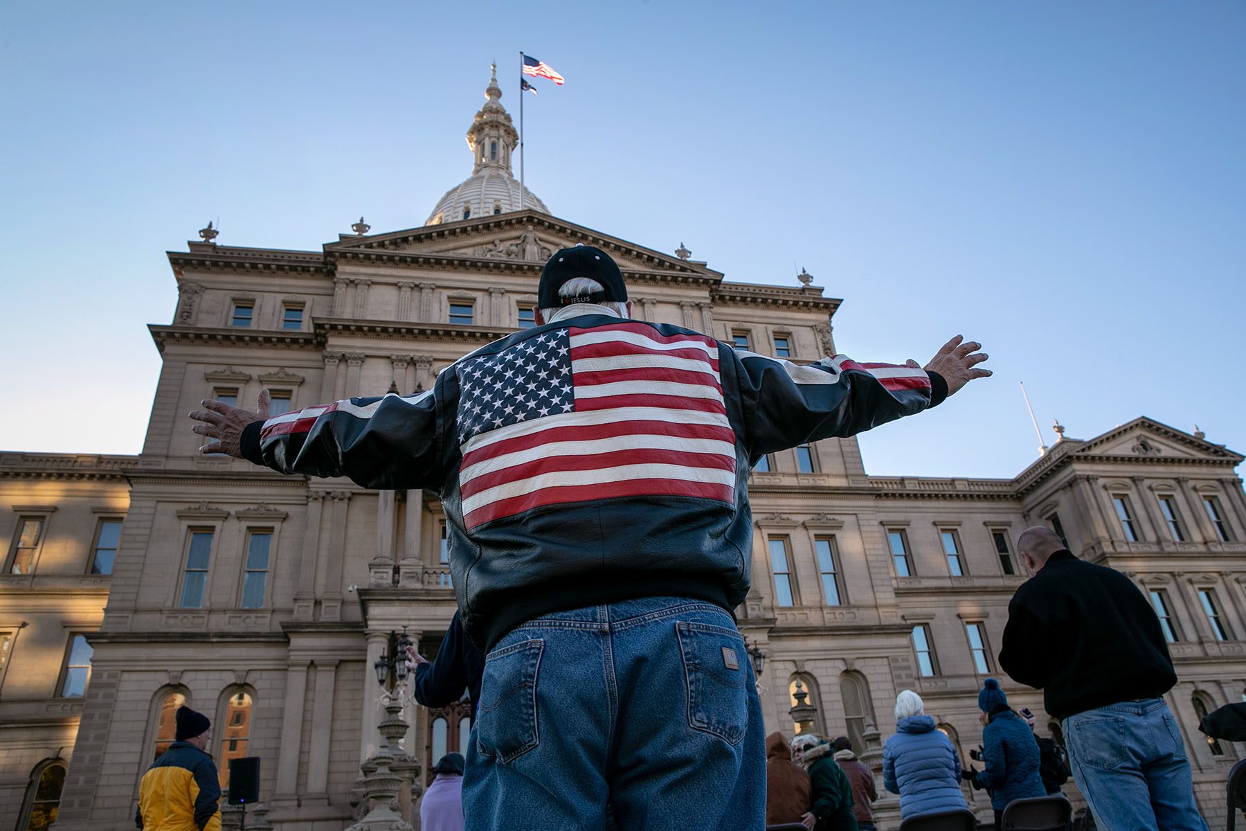 A man wearing a leather jacket featuring an American flag waves his arms as he prays outside the Michigan State Capitol.