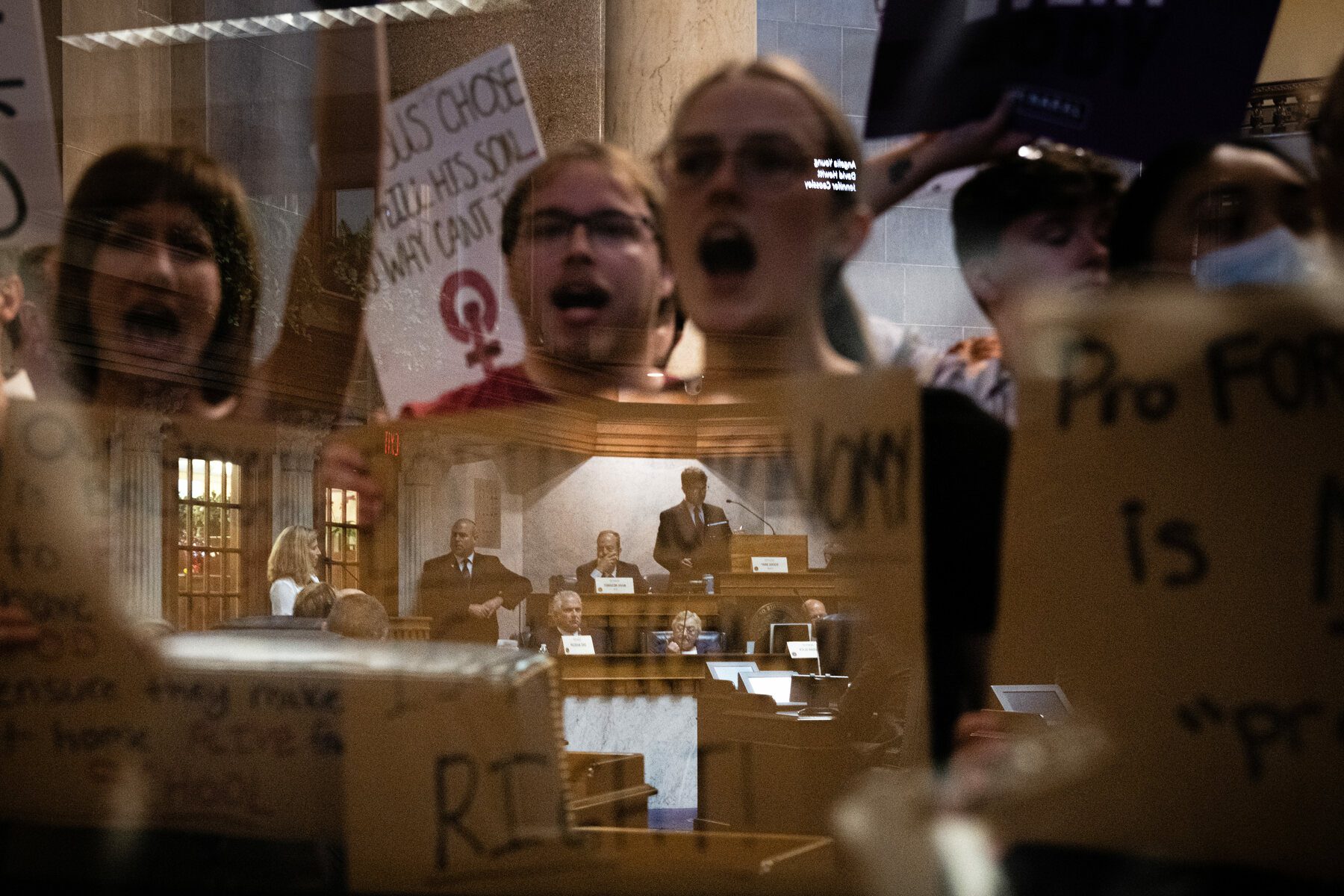 Protestors hold signs and look through a viewing window at the Indiana Capitol building.