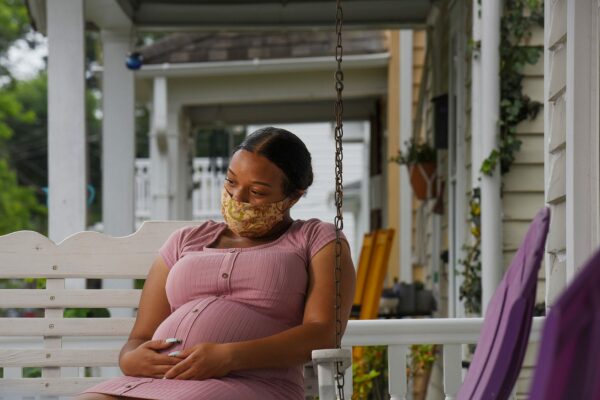 A pregnant woman wearing a face mask sits on a swing on her front porch.