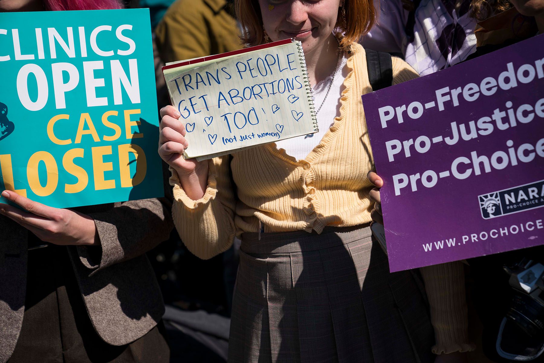 A demonstrator holds up a note that reads "Trans People Get Abortions Too!" (not just women) in the midst of a protest.